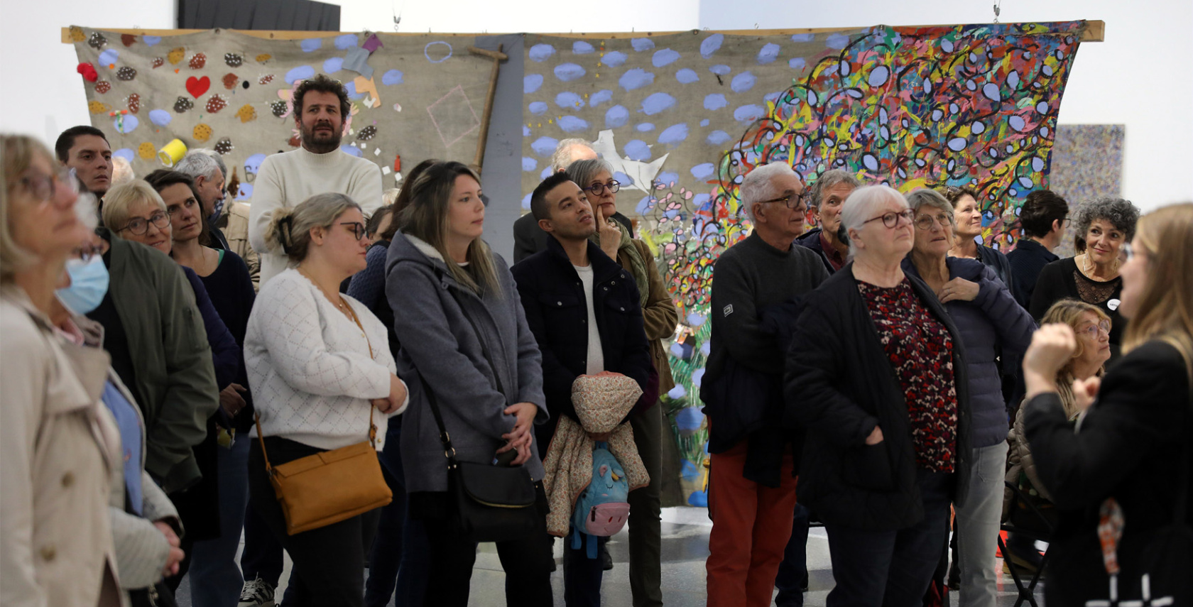 L'image montre une visite commentée avec un groupe de personnes se tenant dos une grande œuvre d'art colorée. L'œuvre est une tapisserie peinte avec de nombreux motifs et couleurs vives, installée au centre de la pièce. Le groupe est composé d'adultes, et ils semblent observer une œuvre hors-champ et la médiatrice avec intérêt. Certains portent des masques.