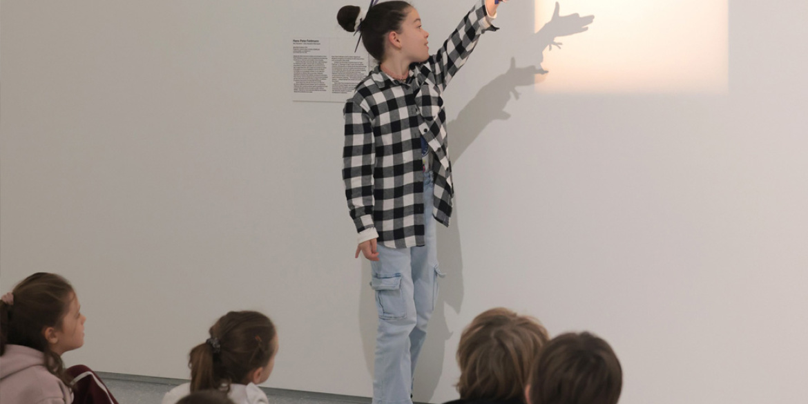 Enfant jouant avec la lumière d'une œuvre d'art, se tenant devant un groupe d'enfants assis. 